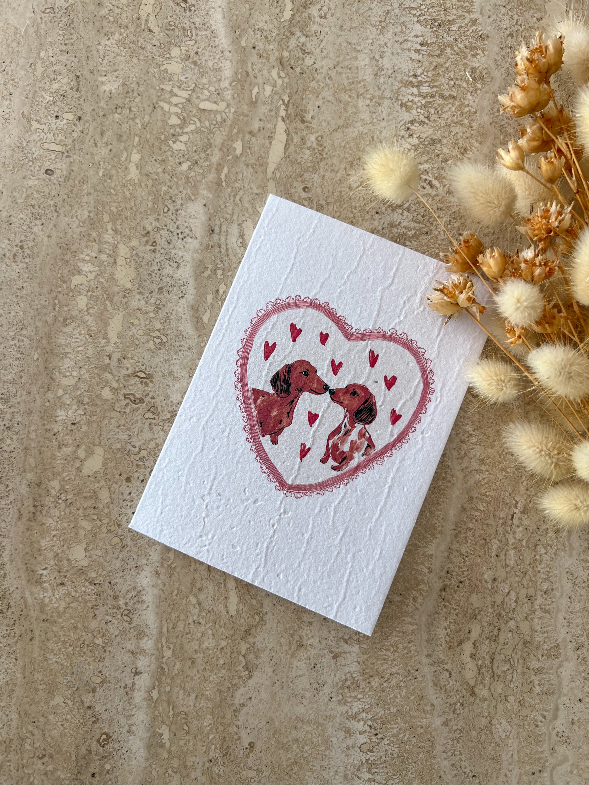 Card with a heart design featuring two dogs and small hearts on a textured surface with dried flowers, by Willow & Sage Co.