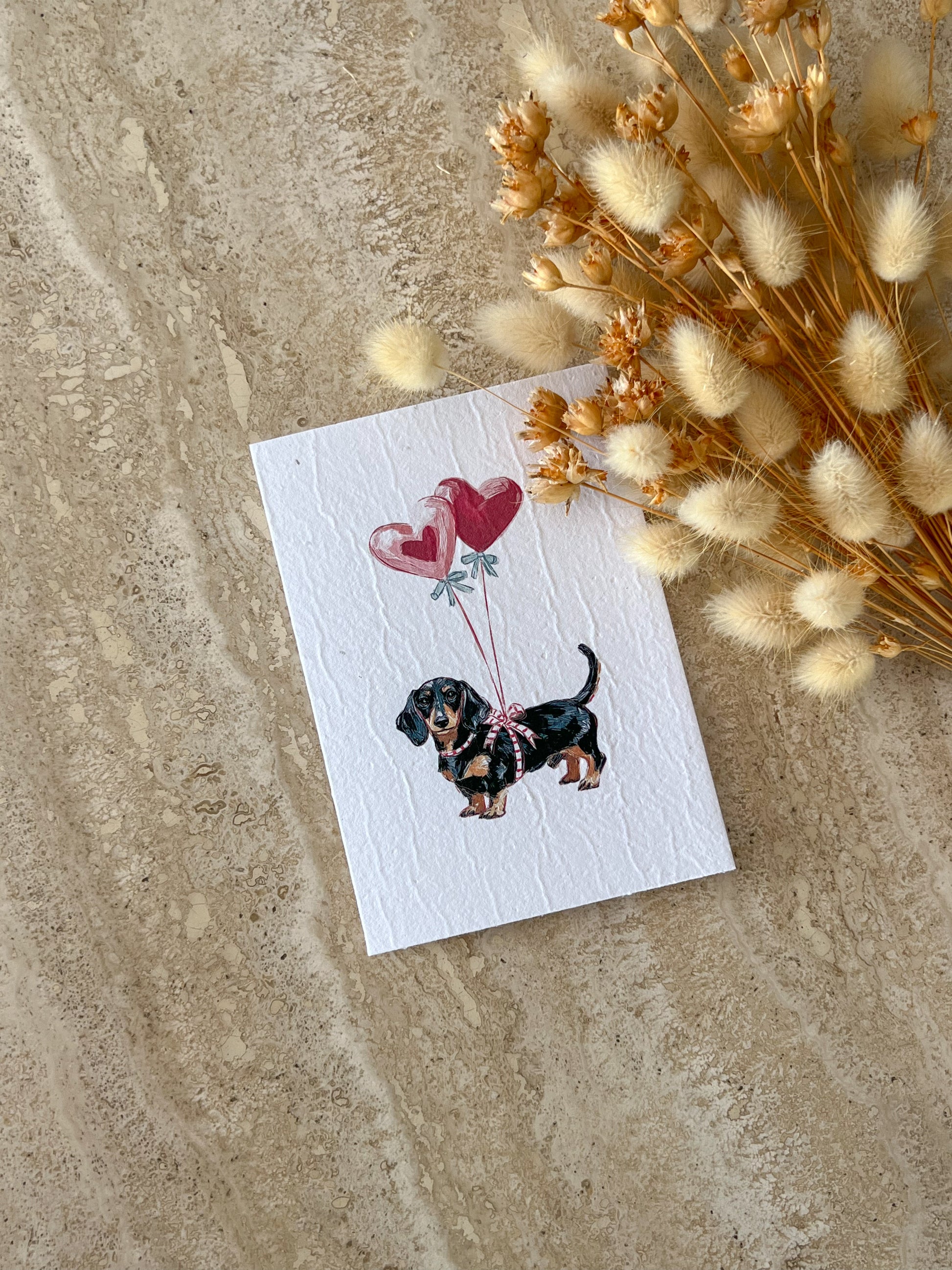 Greeting card with a dachshund illustration holding heart-shaped balloons on a textured surface with dried flowers, by Willow & Sage Co.