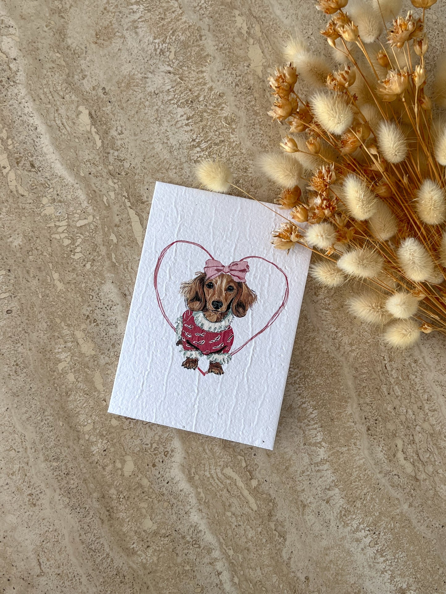 Card with a dachshund dog illustration wearing a pink dress and bow, surrounded by dried flowers on a textured surface, by Willow & Sage Co.