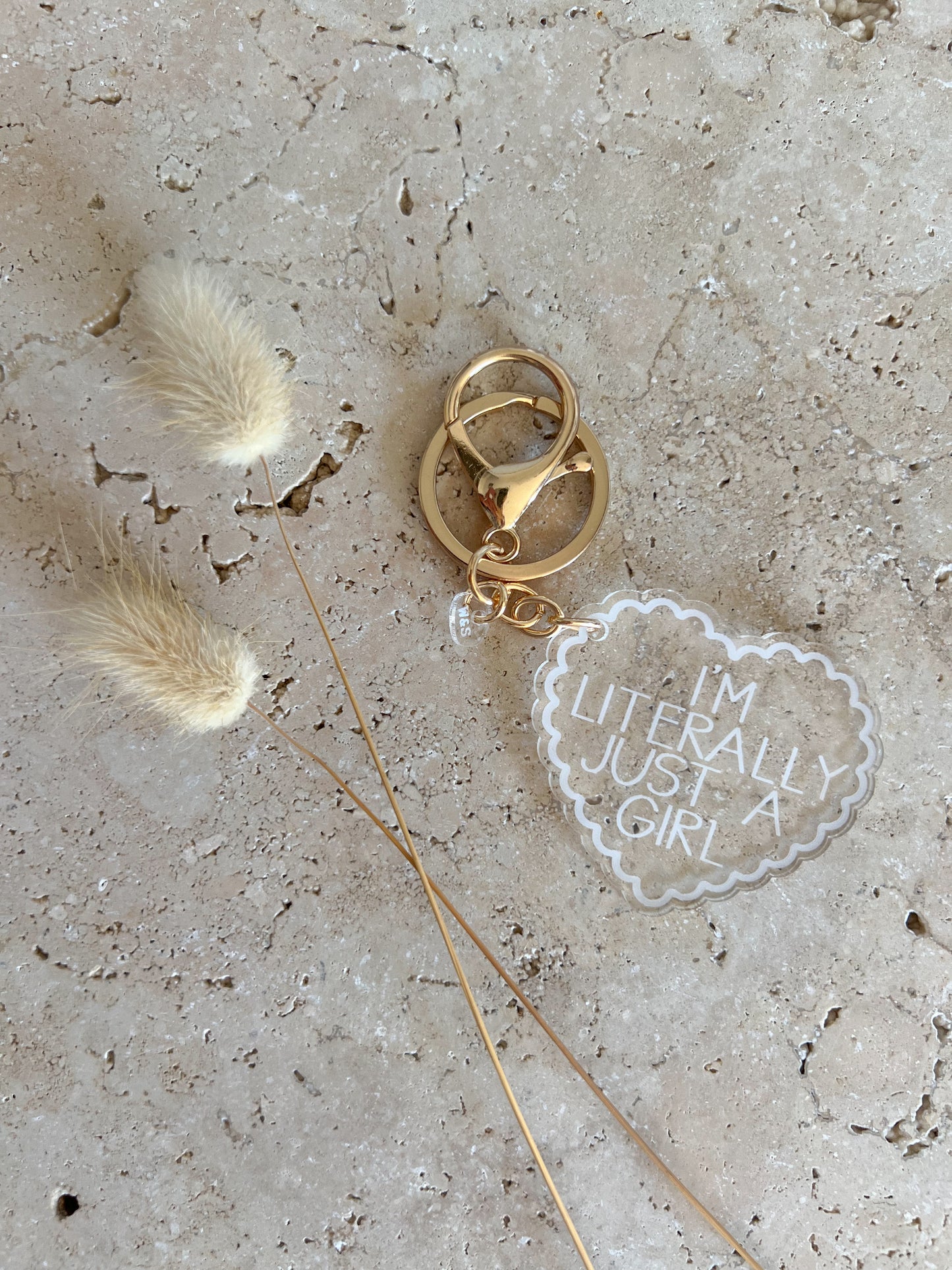 Gold keychain with 'I'm literally just a girl' charm on a textured surface with feathers.