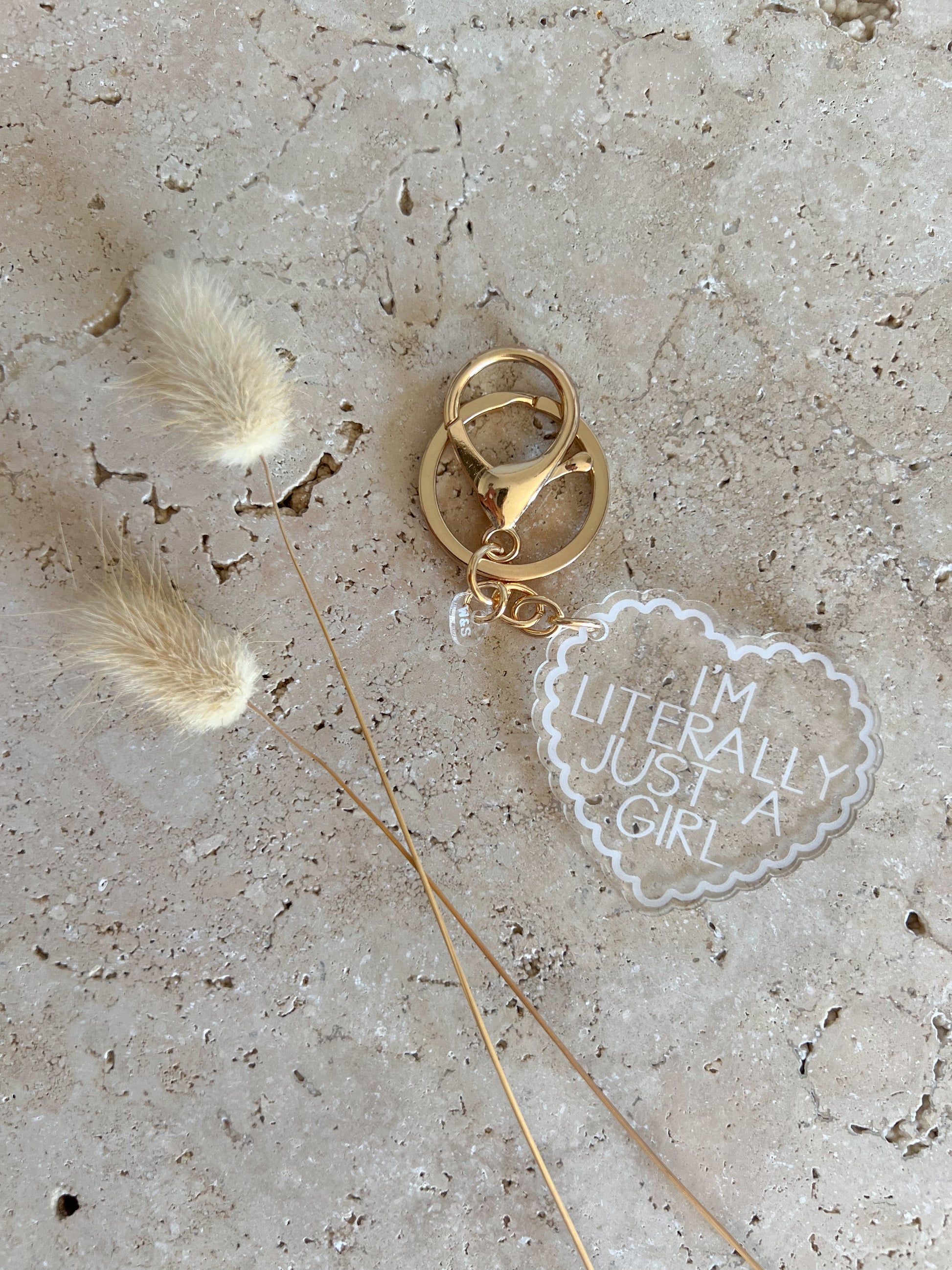 Gold keychain with 'I'm literally just a girl' charm on a textured surface with feathers.