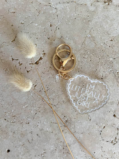 Gold keychain with 'I'm literally just a girl' charm on a textured surface with feathers.