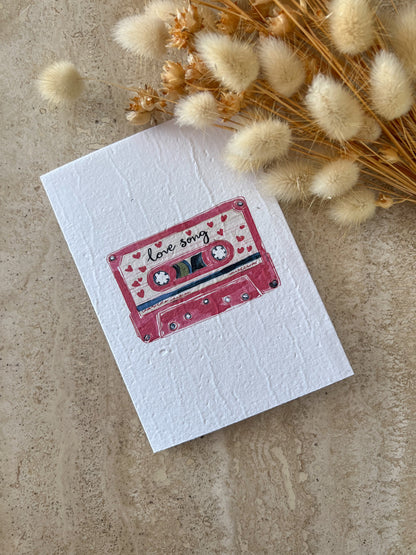 Card with a cassette tape design labeled 'love song' on a textured surface with dried flowers by Willow & Sage Co.