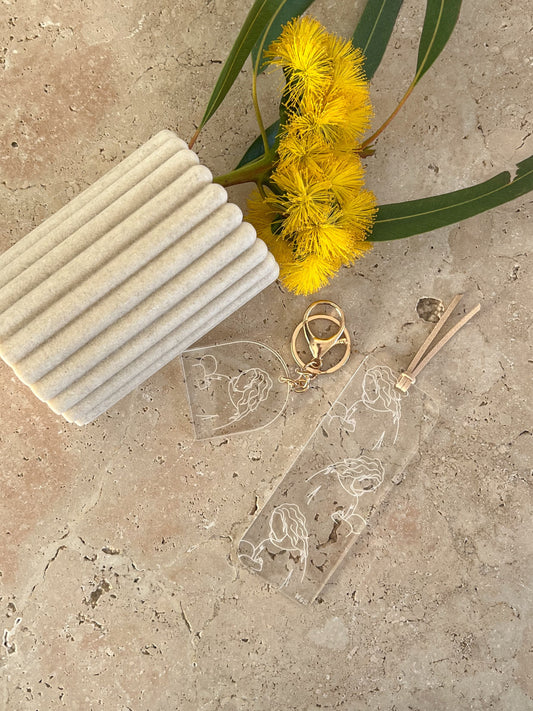 An acrylic bookmark with a beige color, photographed on a textured background alongside a yellow flower and a cord. By willow & sage co